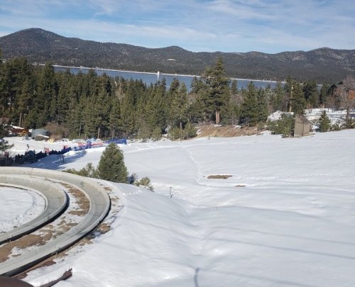 A snowy landscape with trees, a lake in the background, and mountains under a clear blue sky.