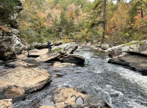 A person stands on a rock by a flowing river, surrounded by autumn-colored trees and rocky terrain.