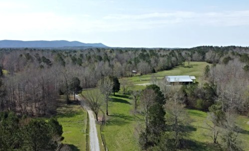 Aerial view of a rural landscape with a winding road, fields, and a barn surrounded by trees and mountains in the distance.
