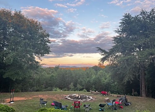 A serene outdoor scene with a fire pit surrounded by chairs, overlooking a forested landscape at sunset.