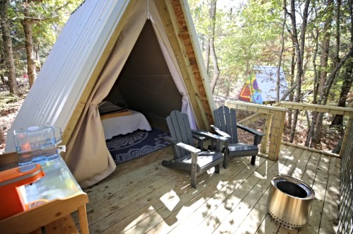 A cozy glamping tent with a wooden deck, two chairs, and a fire pit, surrounded by trees in a serene outdoor setting.