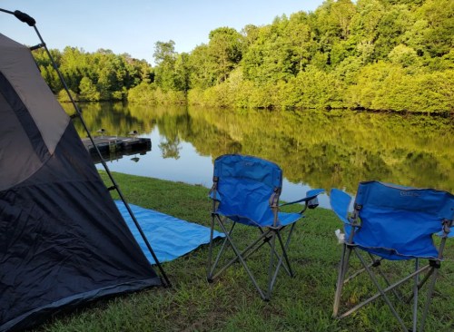 A peaceful campsite by a calm lake, featuring a tent and two blue folding chairs on green grass.