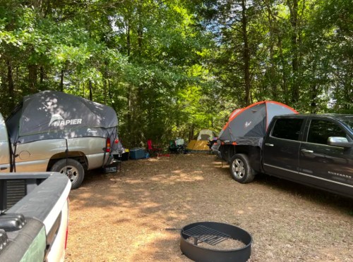 Two trucks with tents set up in a wooded campsite, surrounded by trees and camping gear.