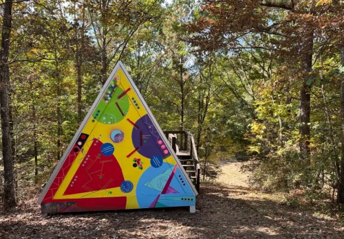 A colorful triangular structure in a wooded area, surrounded by trees and a path leading into the forest.