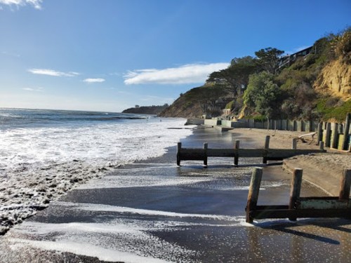 A serene beach scene with gentle waves, wooden posts, and a coastal cliff under a clear blue sky.