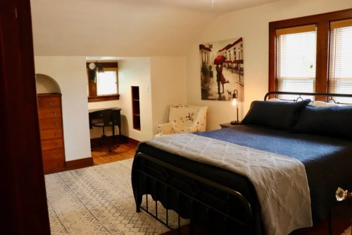 Cozy bedroom with a metal bed, desk, chair, and artwork, featuring warm lighting and wooden accents.