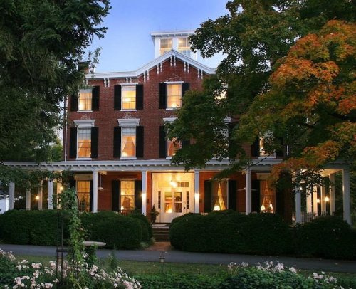 A large brick house with white trim, surrounded by greenery and trees, illuminated at dusk.