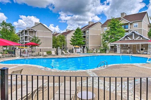 A sunny pool area with lounge chairs, surrounded by trees and apartment buildings, under a partly cloudy sky.