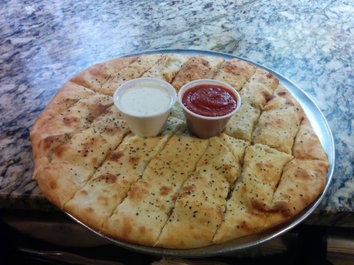 A round platter of cheesy breadsticks, cut into pieces, served with marinara and ranch dipping sauces.