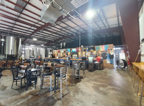 Interior of a brewery with metal brewing tanks, wooden tables, and patrons enjoying drinks in a spacious setting.