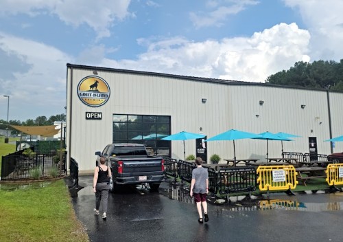 Two people walk towards a building with a sign that says "Goat Island" and outdoor seating under blue umbrellas.