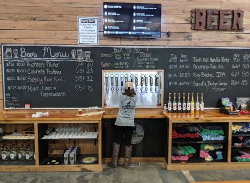 A person pours beer from a tap at a bar with a chalkboard menu and wooden counters displaying various drinks.