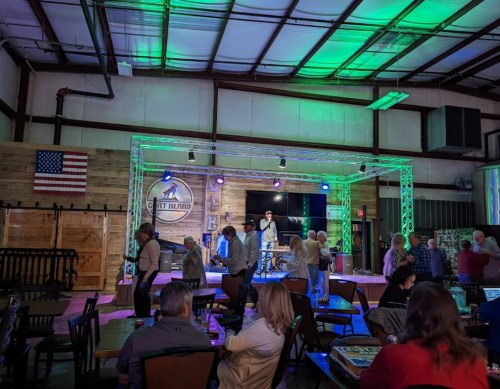 A lively indoor venue with a stage, colorful lights, and people socializing at tables. An American flag is displayed.
