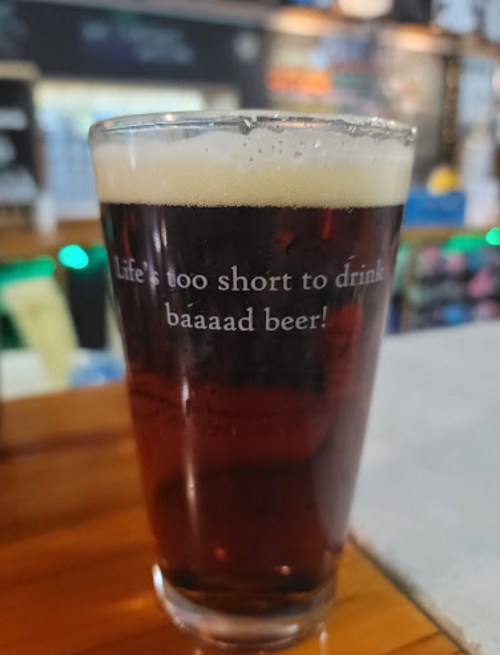 A pint of dark beer with a humorous quote: "Life's too short to drink baaaad beer!" in a bar setting.