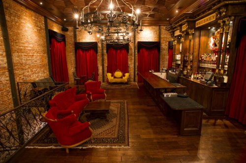 A cozy bar with red velvet curtains, wooden floors, and a stylish seating area featuring red chairs and a dark wood bar.
