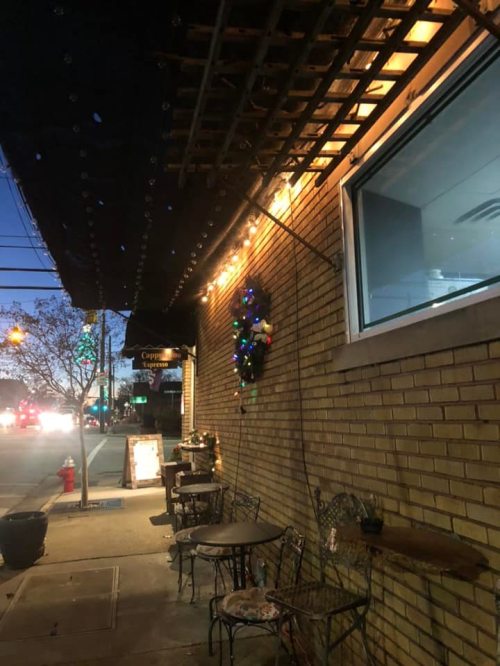 A cozy street scene at dusk, featuring a brick wall adorned with lights and a festive wreath, with outdoor seating nearby.