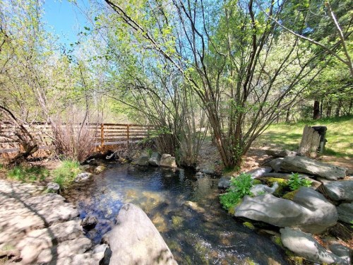 A serene stream flows through a lush green landscape, surrounded by trees and rocks, with a wooden fence in the background.
