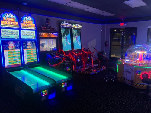 A brightly lit arcade featuring various games, including lane master and racing simulators, in a darkened room.