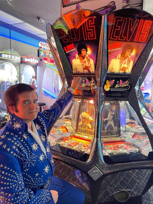 A performer in a blue sequined suit poses next to an Elvis-themed arcade machine in a vibrant game room.