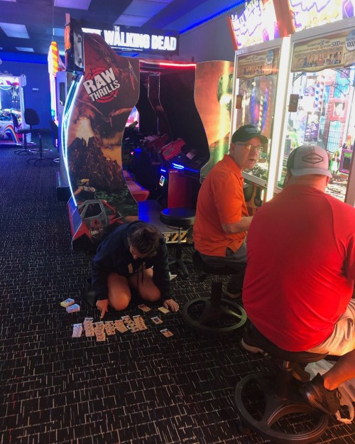 A girl sits on the floor organizing tickets in an arcade, while two men play games at nearby machines.