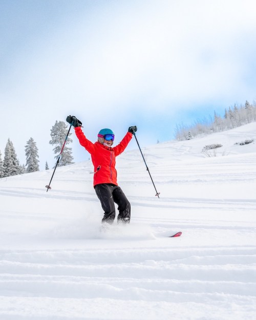 A skier in a red jacket and blue helmet joyfully skiing down a snowy slope with arms raised.