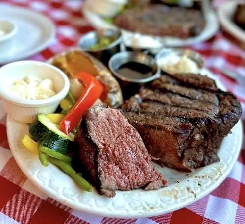 A plate of grilled steak with sides of vegetables, baked potato, and dipping sauces on a checkered tablecloth.