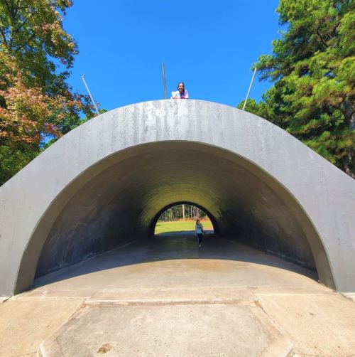 A person stands on top of a large, curved structure, while another walks through the arch below, surrounded by trees.