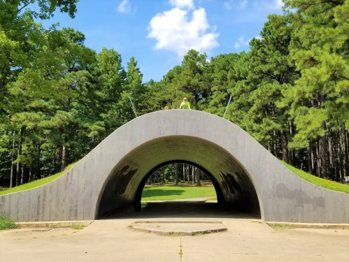 A curved concrete structure in a wooded area, with a person standing on top under a blue sky with clouds.