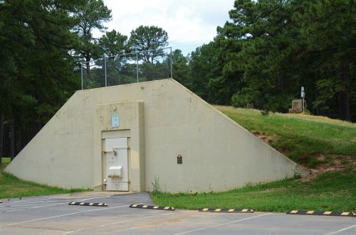 A concrete bunker with a sloped roof, surrounded by trees and a fenced area, located in a parking lot.