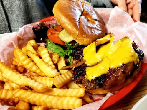 A juicy cheeseburger with lettuce, tomato, and pickles, served with crinkle-cut fries in a red basket.