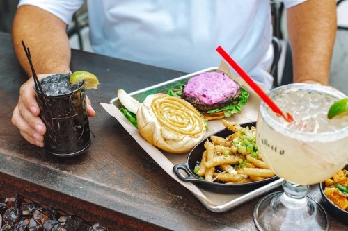 A person holds a cocktail next to a tray with a burger, fries, and a cinnamon roll.