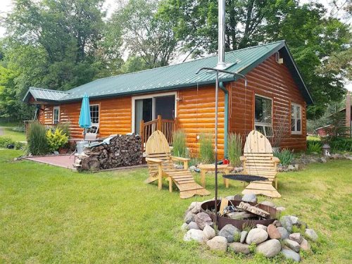 A cozy log cabin with a green metal roof, surrounded by greenery and a fire pit with wooden chairs in the foreground.