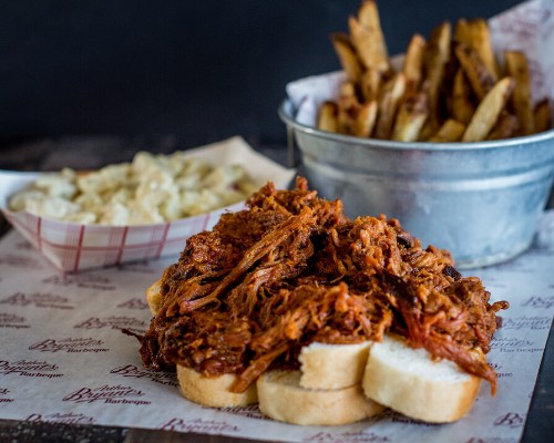 A plate of pulled pork on bread, served with fries in a bucket and a side of macaroni and cheese.