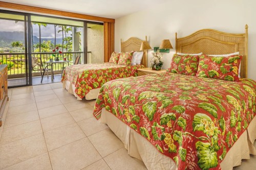 Bright hotel room with two beds, tropical-patterned bedding, and a balcony overlooking a scenic view.
