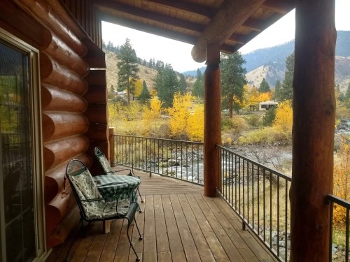 A cozy log cabin porch with two chairs overlooking a scenic view of autumn trees and a river.