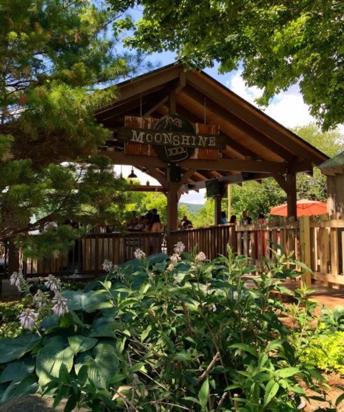 A wooden pavilion labeled "Moonshine" surrounded by greenery, with people enjoying the outdoor space.