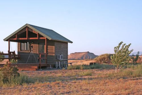 A small wooden cabin in a grassy area with a clear sky and distant hills in the background.