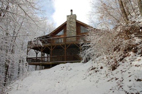 A cozy wooden cabin with a stone chimney, surrounded by snow-covered trees and a winter landscape.