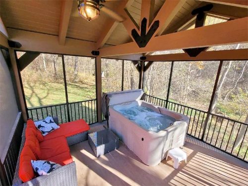 A screened porch featuring a hot tub, a red sofa, and a small table, surrounded by trees and greenery.
