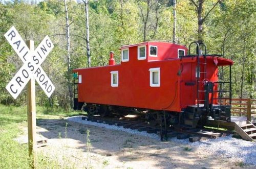 A red caboose parked near a railroad crossing sign, surrounded by trees and gravel.
