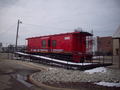 A red train caboose from 1883, displayed on a raised platform with a gravel area and cloudy sky in the background.