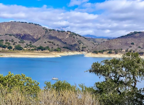 A serene lake surrounded by rolling hills and sparse vegetation under a partly cloudy sky. A small boat is visible on the water.