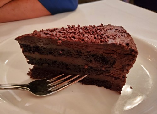 A slice of rich chocolate cake with chocolate shavings on top, served on a white plate with a fork beside it.