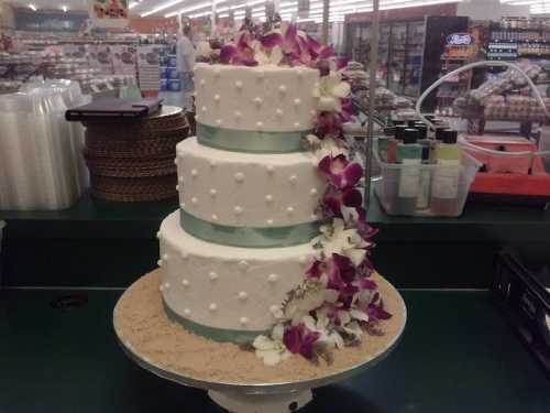 A three-tiered white wedding cake adorned with purple orchids and a light blue ribbon, resting on a sandy base.