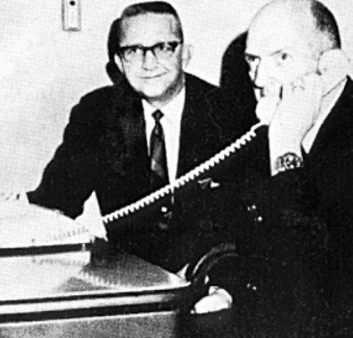 Two men sit at a desk, one on the phone, smiling at the camera in a vintage black-and-white photo.