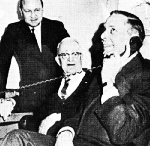 Three men in suits, one on the phone, smiling and engaged in conversation in a vintage office setting.