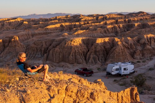 A person relaxes in a chair on a rocky ledge, overlooking a desert landscape with a camper and truck below.