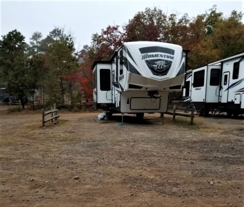 A large RV parked in a wooded area, surrounded by trees with autumn foliage. Other RVs are visible in the background.