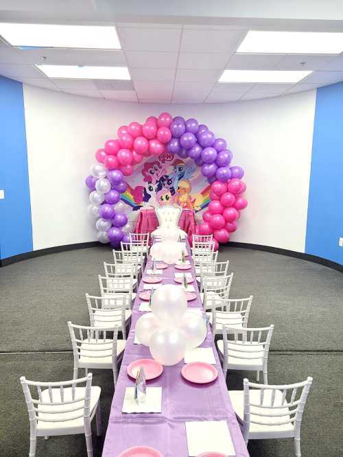 A decorated party space with a long table, pink and purple balloons, and a colorful backdrop for a celebration.