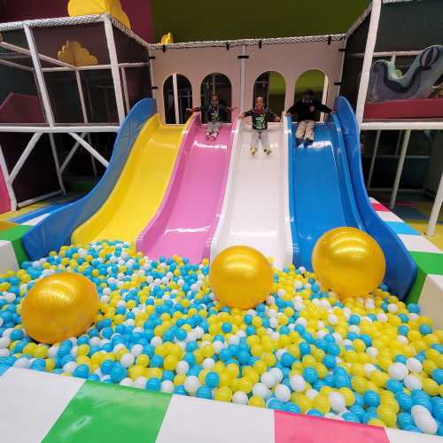 Three children sit on colorful slides above a ball pit filled with blue and yellow balls, with large golden balls nearby.